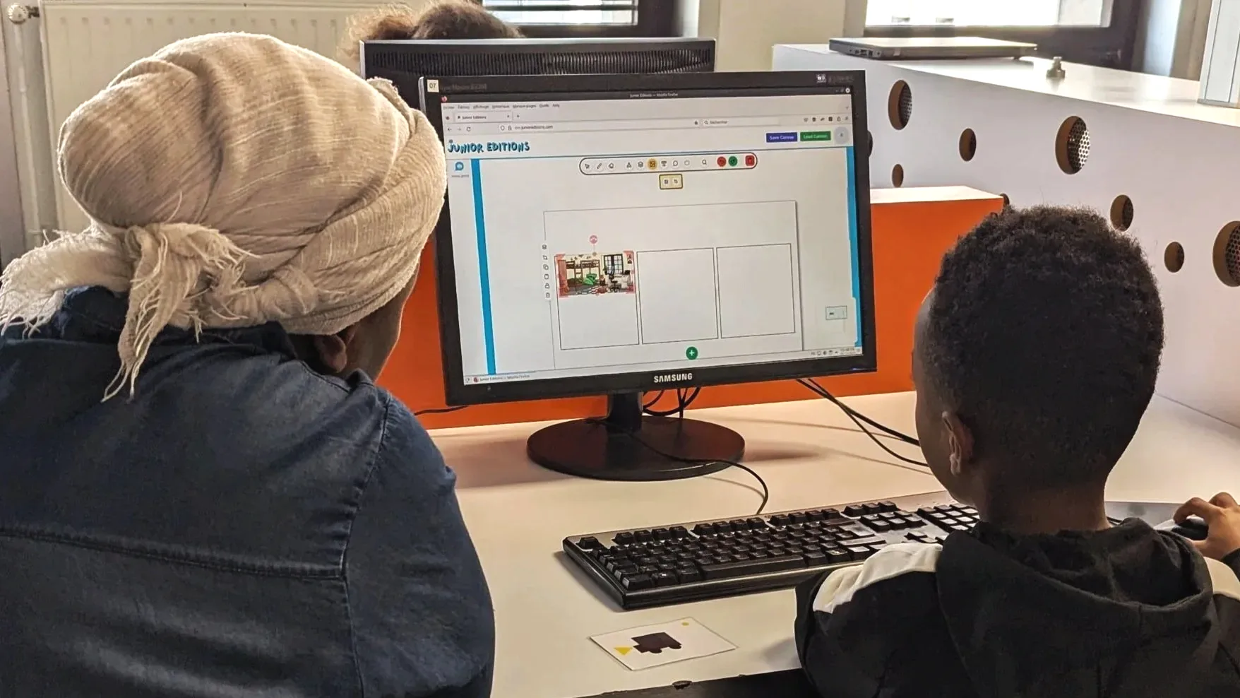 A mother and her son work together at a desk computer, creating a layout for a book or publication, in a computer room of the library.