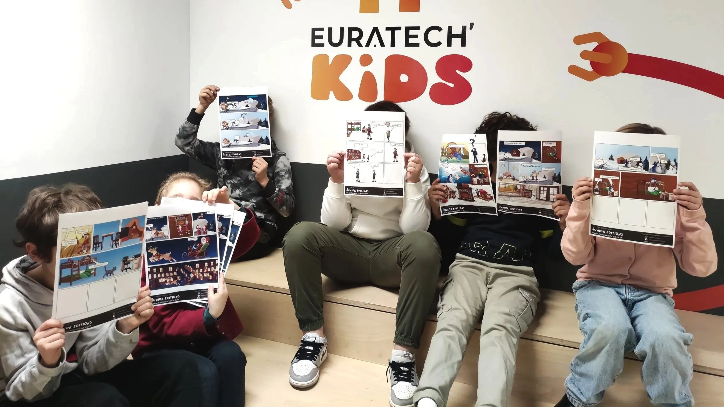 A group of children sitting against a white wall with an 'EURATECH KIDS' poster in the background, each holding a printed comic sheet with various scenes.