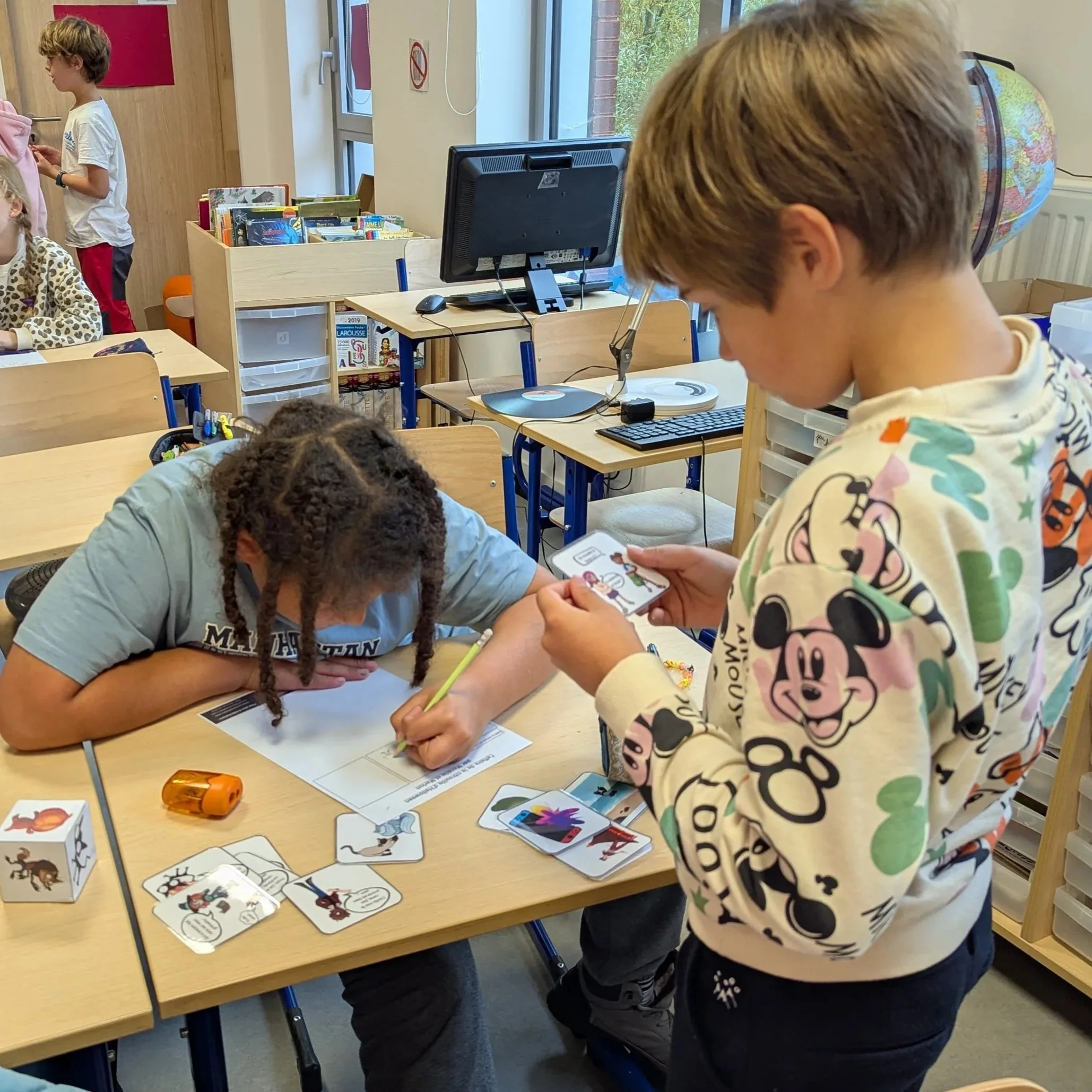 A classroom with children playing with cards illustrated with different creatures and characters, called writing starters. Some children are sitting at a table or standing, concentrating or interacting.