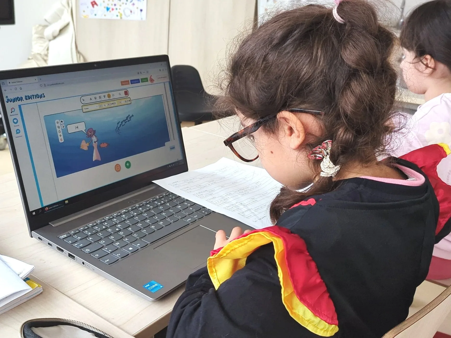 A young girl wearing a red, yellow and black scarf works in front of a laptop in a classroom. The computer screen shows an educational website for children.