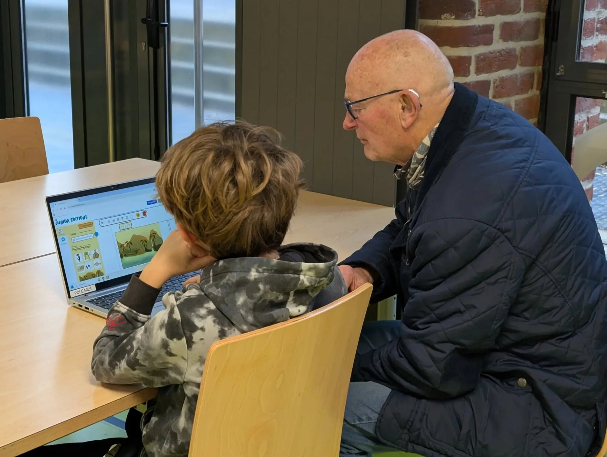 A grandfather and his grandson sitting at a table, looking at a laptop indoors, in the middle of creating a digital comic