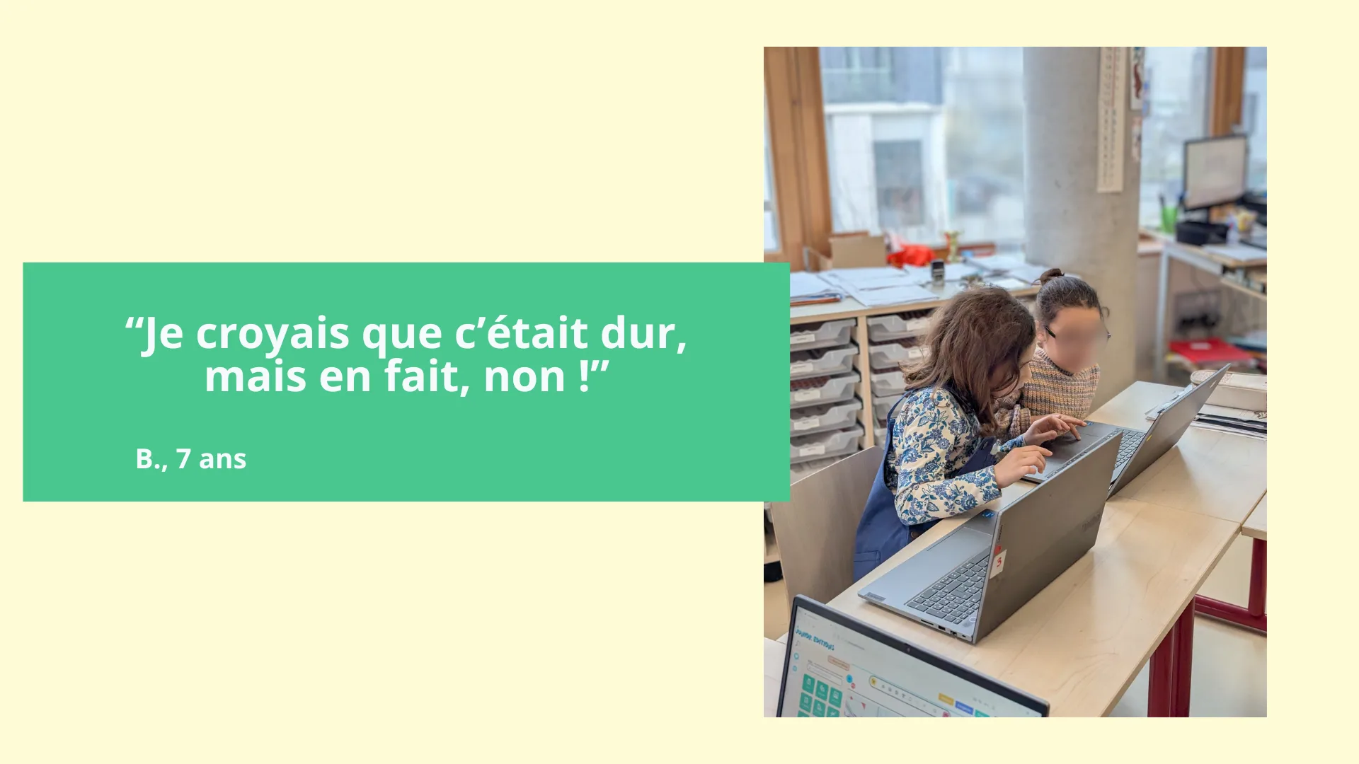 Two children sitting at a school table work on laptops in a classroom. The room has large windows and storage units with drawers. One of the children wears a long-sleeved sweatshirt with blue patterns.