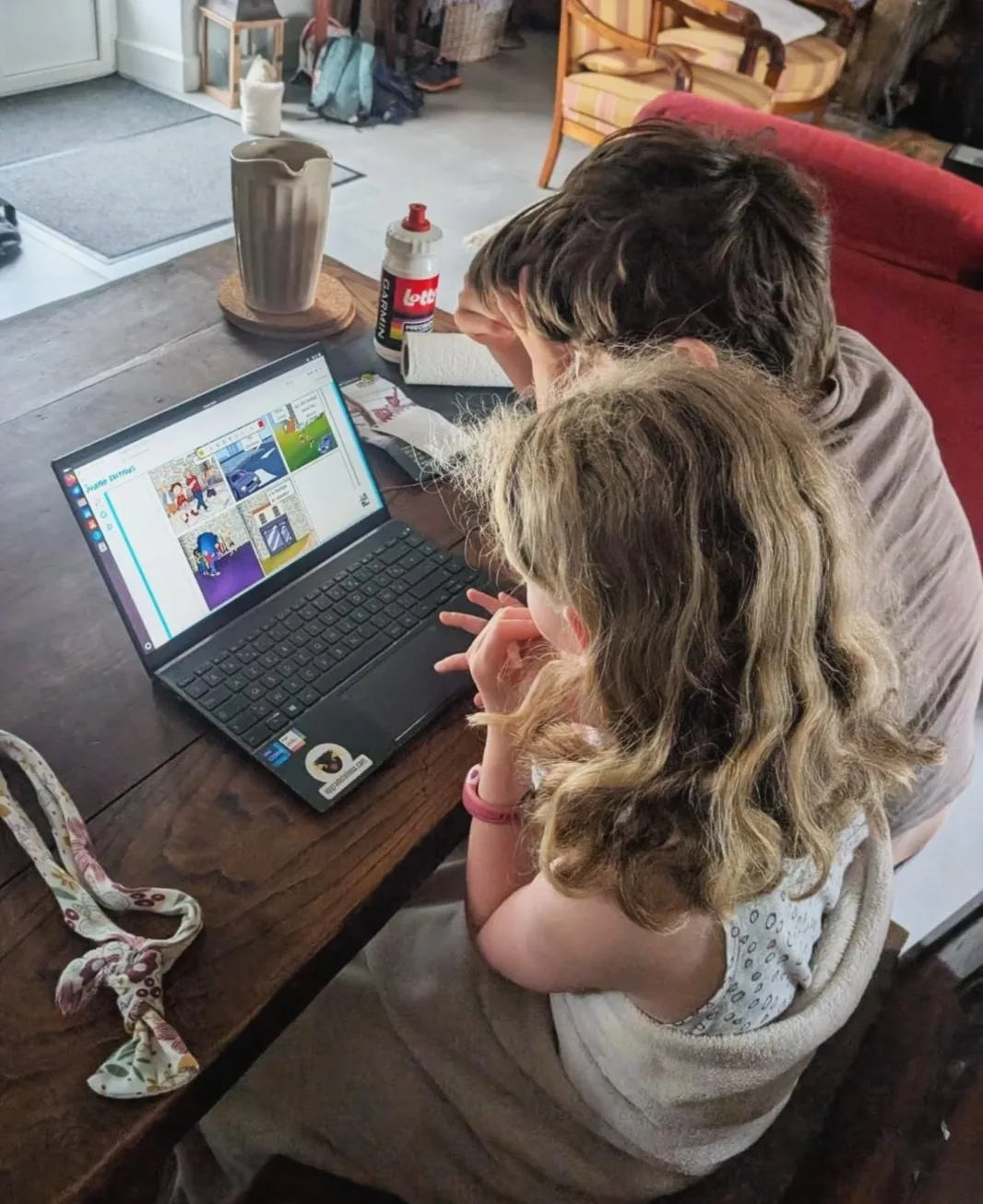 Two children look at a laptop on a wooden table, in a bright room with furniture and various objects. The scene seems to take place at home, with a comfortable setting.