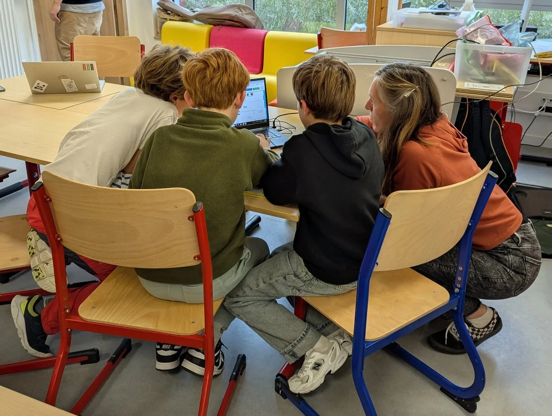 Five young people sitting at a table looking at a laptop together with their teacher. The room is bright with large windows at the back and colourful chairs.